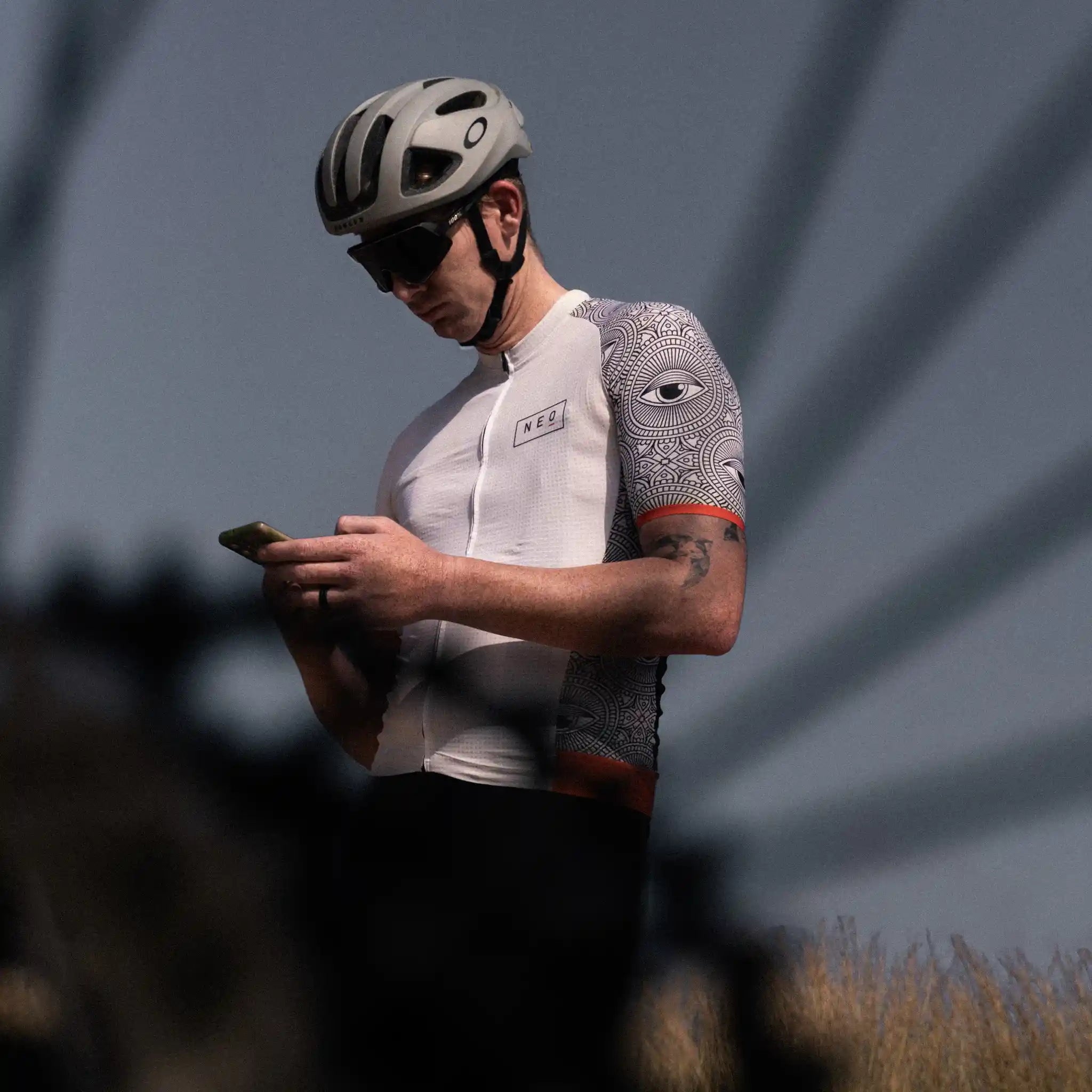 A cyclist wearing a Neo Hyphen 'All-Seeing Eye' cycling jersey looks at his phone, possibly checking directions or Strava.