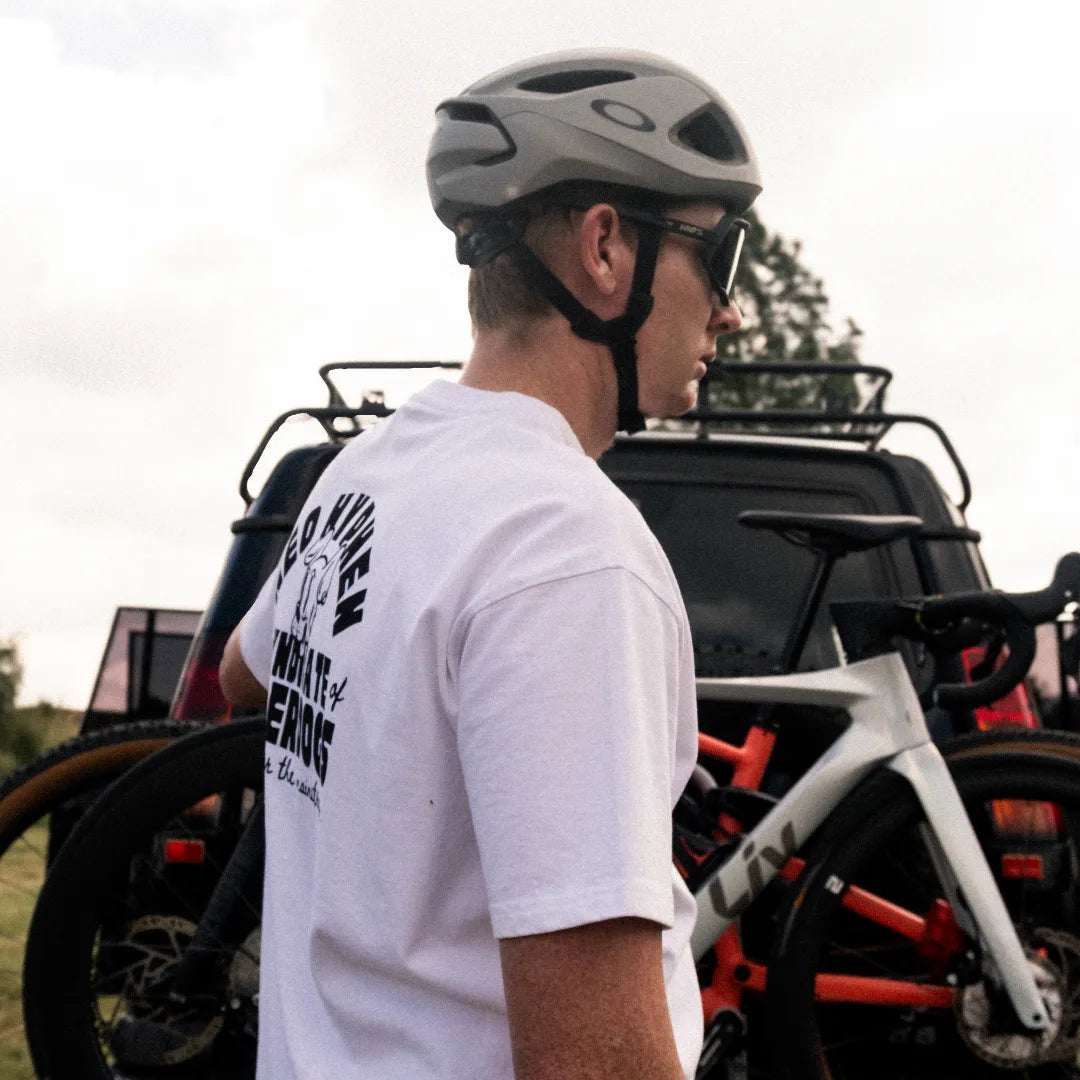 A man in a cycling helmet and a Neo Hyphen white 'Syndicate of the Underdogs' 100% cotton t-shirt stands beside a car, looking relaxed and ready for an adventure.