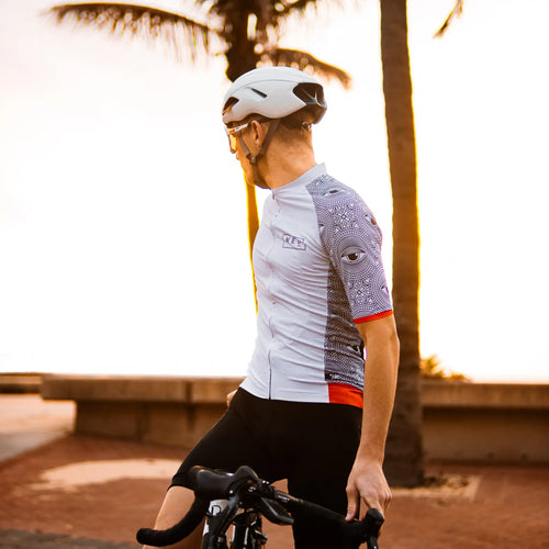 A cyclist in a Neo Hyphen All-Seeing Eye Jersey and helmet poses on a sidewalk, ready for a ride.