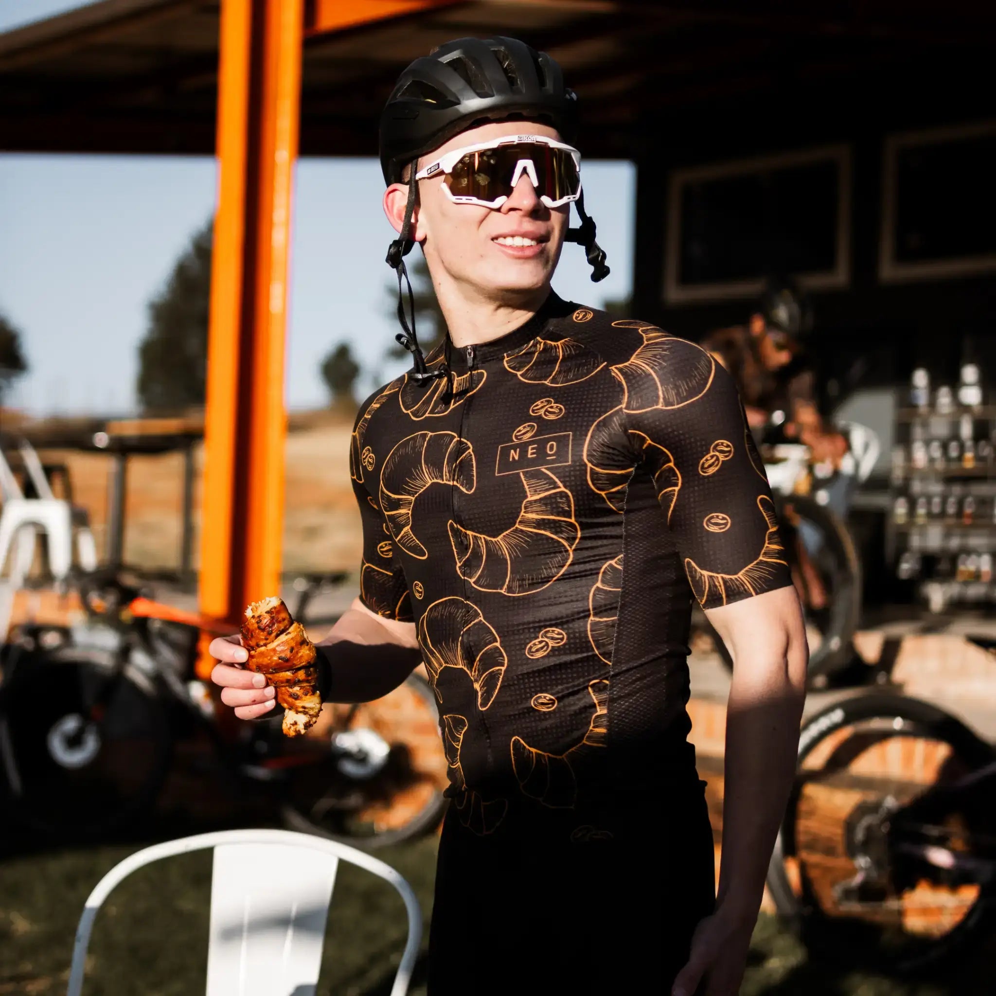 A man with cycling sunglasses and a helmet, styling a Neo Hyphen Le Croissant Jersey, set for a fun ride.