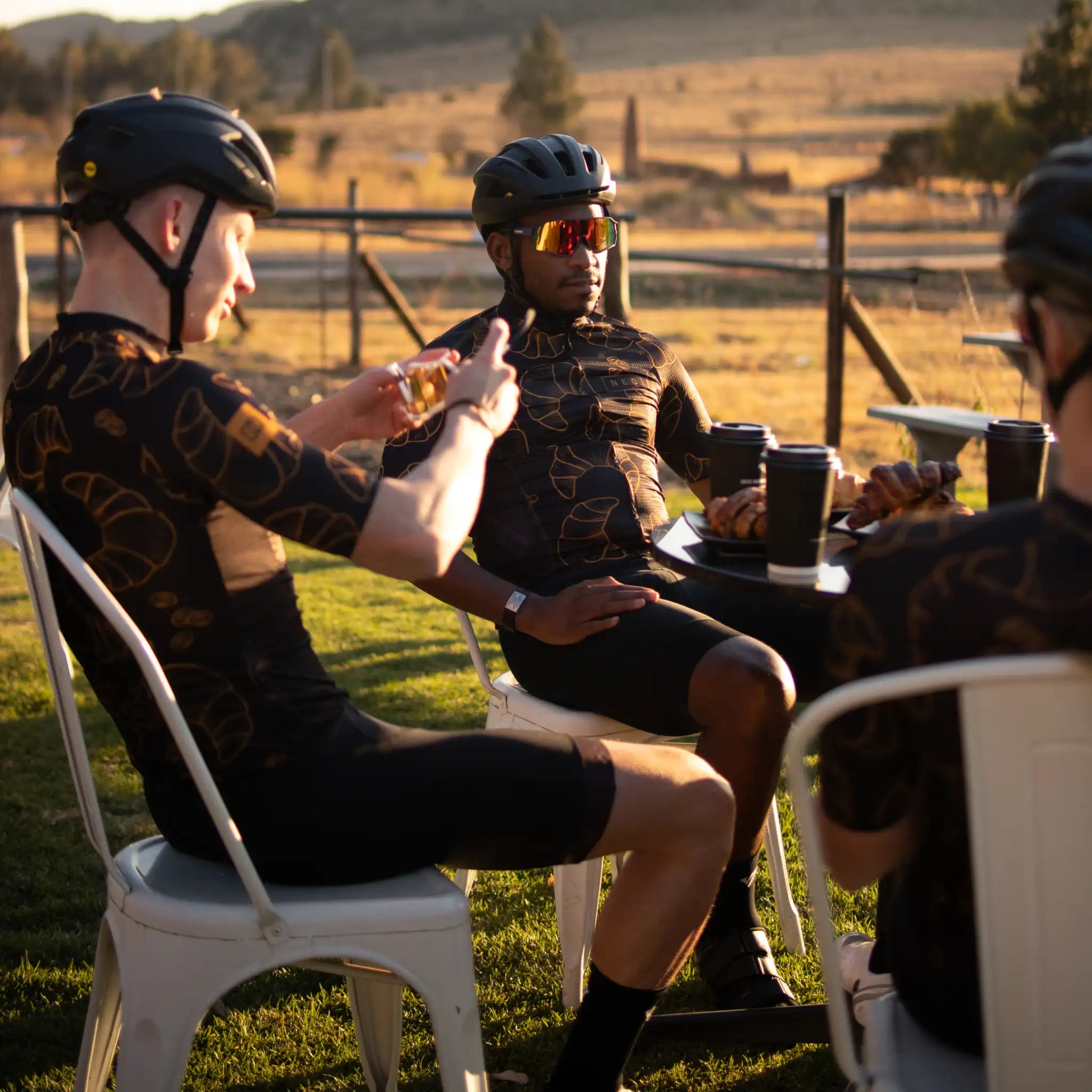A group of male cyclists in chairs enjoying coffee together, wearing Neo Hyphen Le Croissant jerseys.