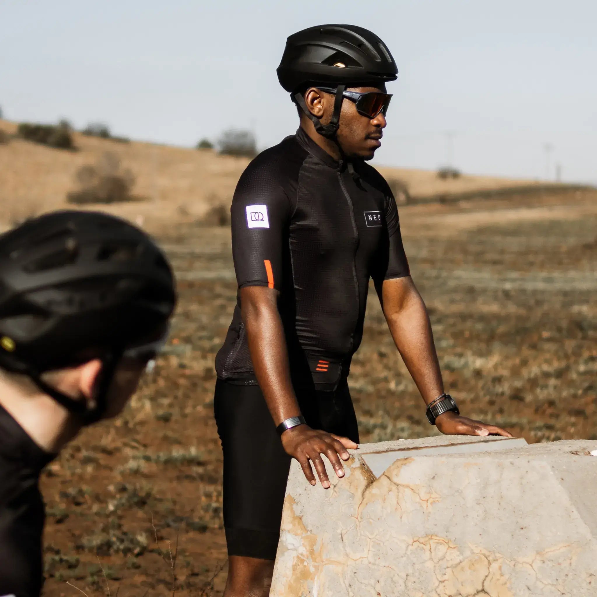 Two cyclists wearing Neo Hyphen Phantom cycling jerseys pose beside a rock.