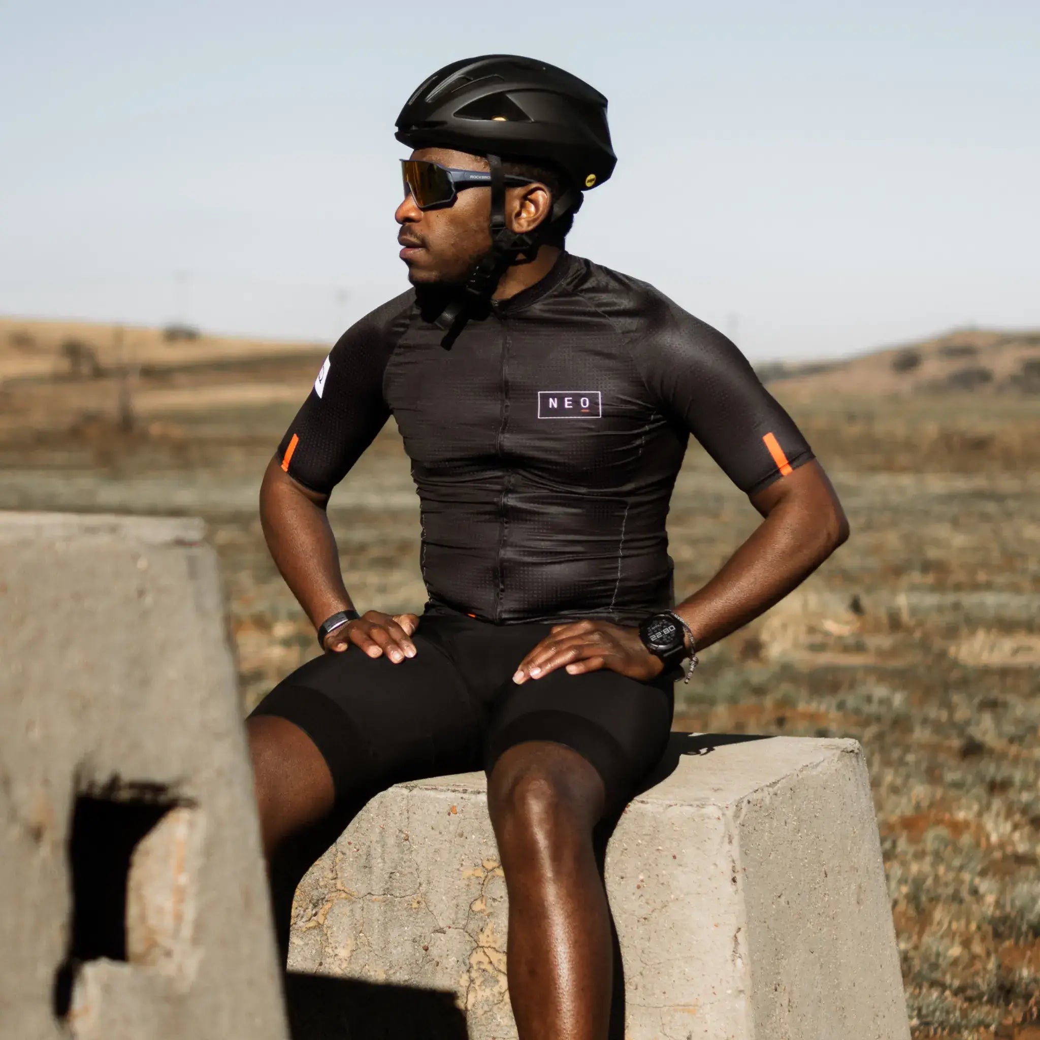 A cyclist wearing a black Neo Hyphen Phantom Jersey and a helmet sits on a cement block, looking relaxed and prepared.