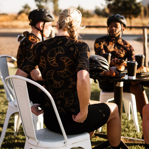 A diverse group of cyclists gathered around a table, enjoying conversation in a cozy setting wearing Neo Hyphen Le Croissant cycling jerseys.
