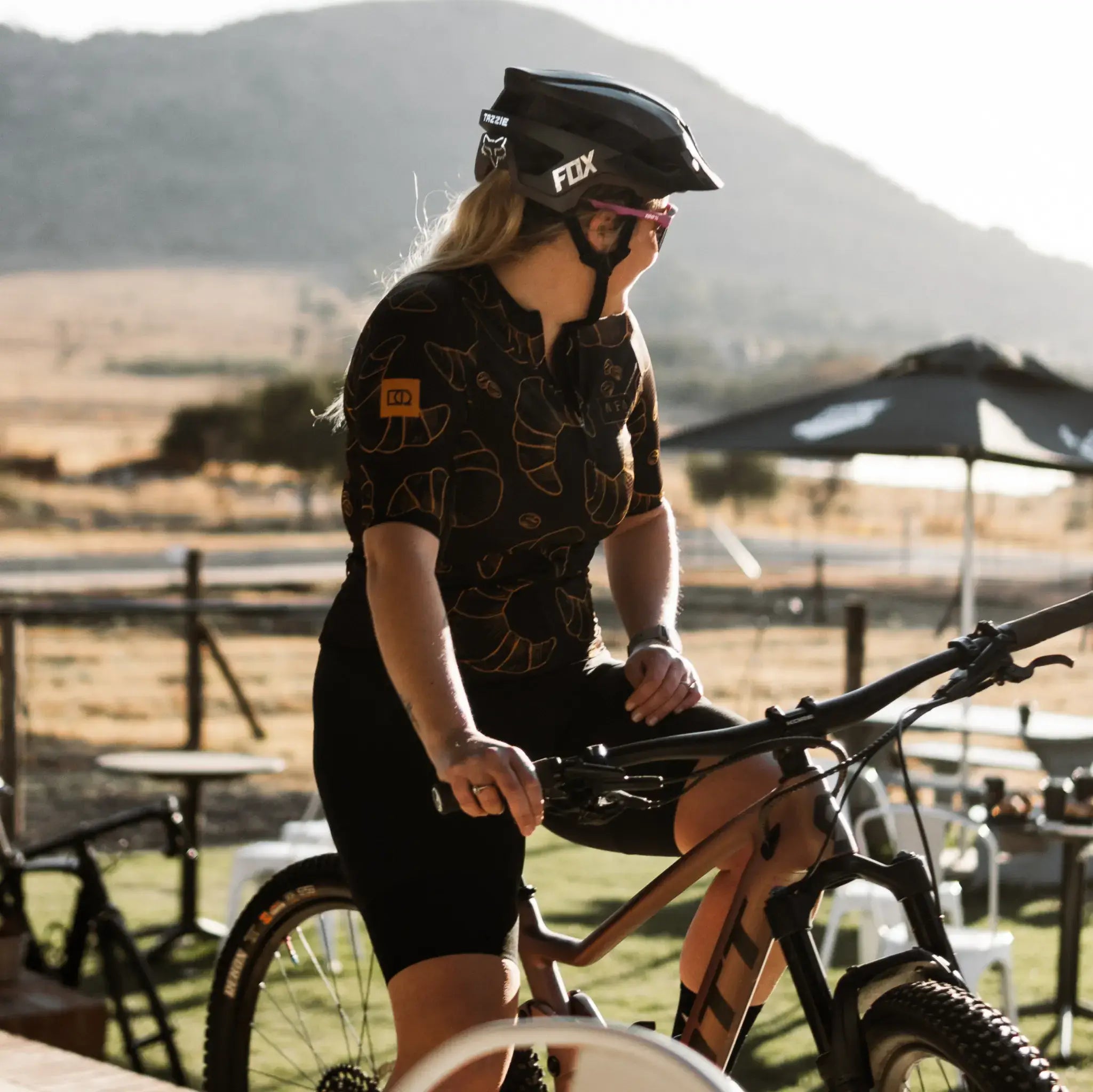 A woman in a helmet sits on her mountain bike, sporting a Neo Hyphen Le Croissant Jersey, prepared for cycling.