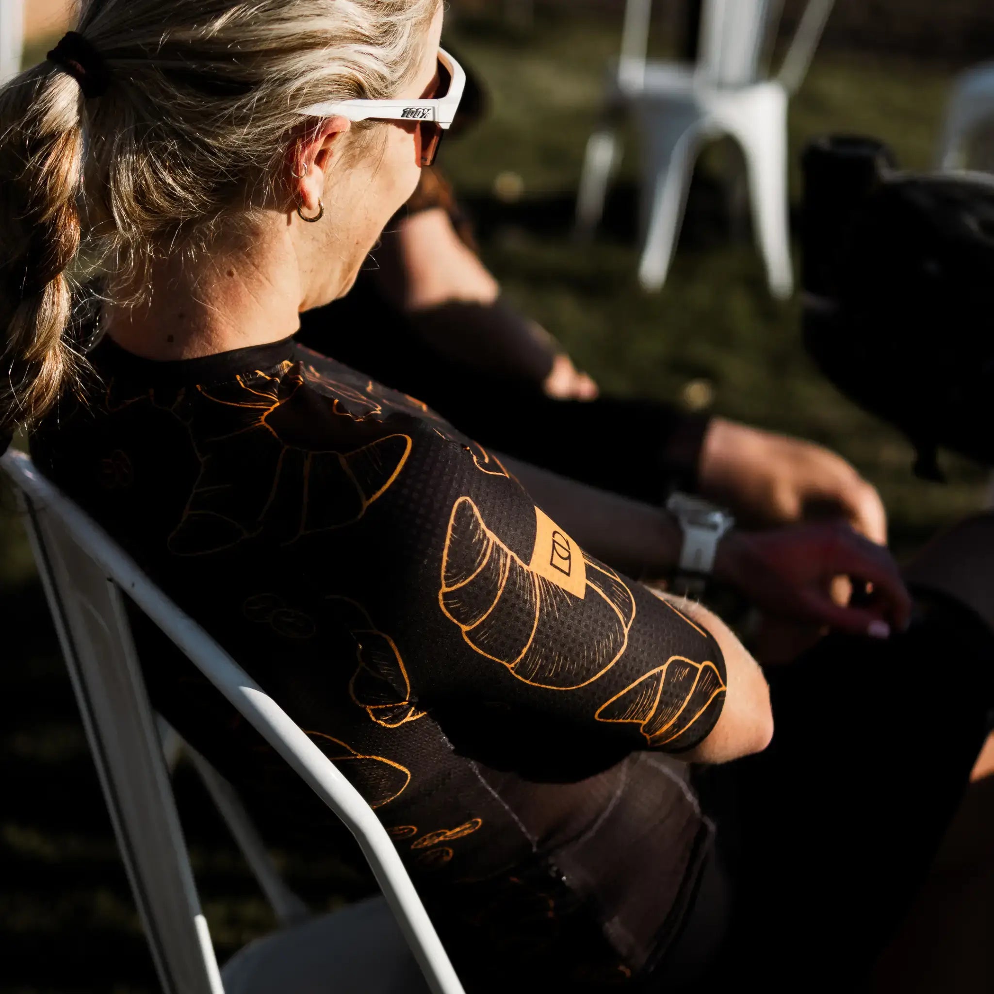 A woman wearing a Neo Hyphen Le Croissant cycling jersey, seated on a chair, looking relaxed.